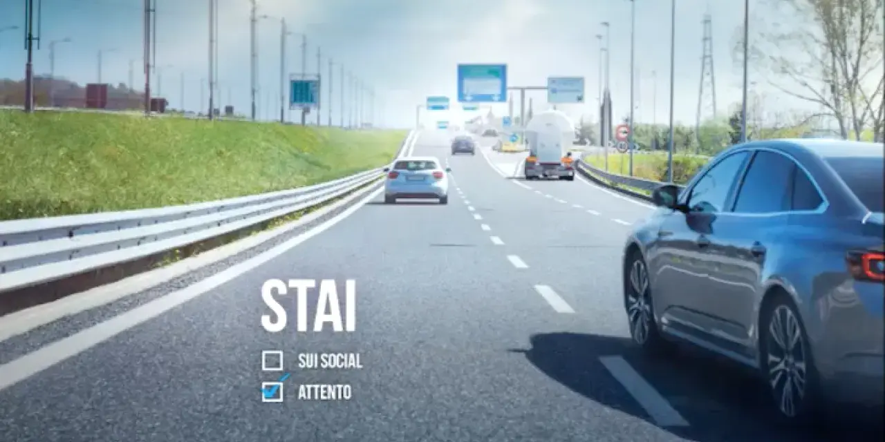 Sulla strada, auto e camion procedono. "STAI ATTENTO" è scritto, ricordando l'art. 79 CDS.
