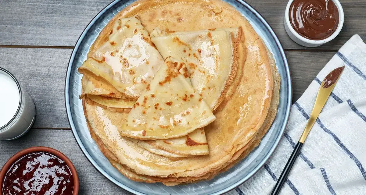Pile de crêpes dorées, parfaites pour une recette pâte à crêpes. Accompagnées de confiture, pâte à tartiner et verre de lait.