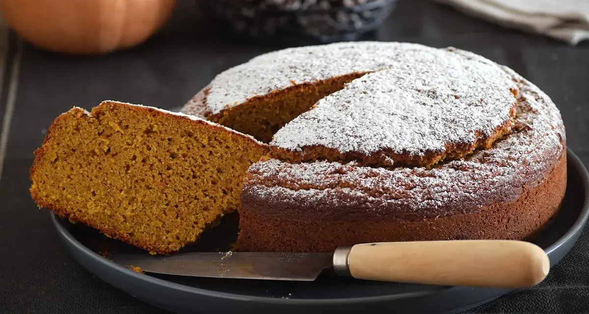 Bizcocho de calabaza esponjoso y dorado, espolvoreado con azúcar glas. Una rebanada muestra su interior húmedo y aromático.