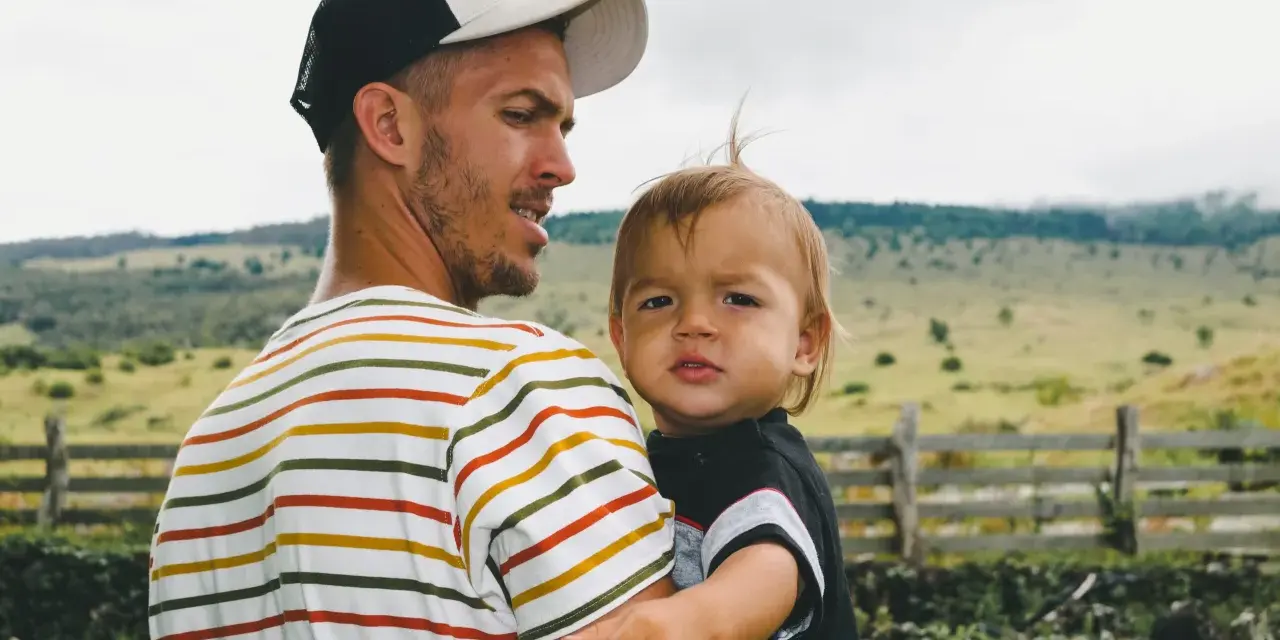 Ein Mann mit Bart und Kappe trägt ein Kind. Die Eigenschaften eines Papas zeigen sich in seiner liebevollen Umarmung.