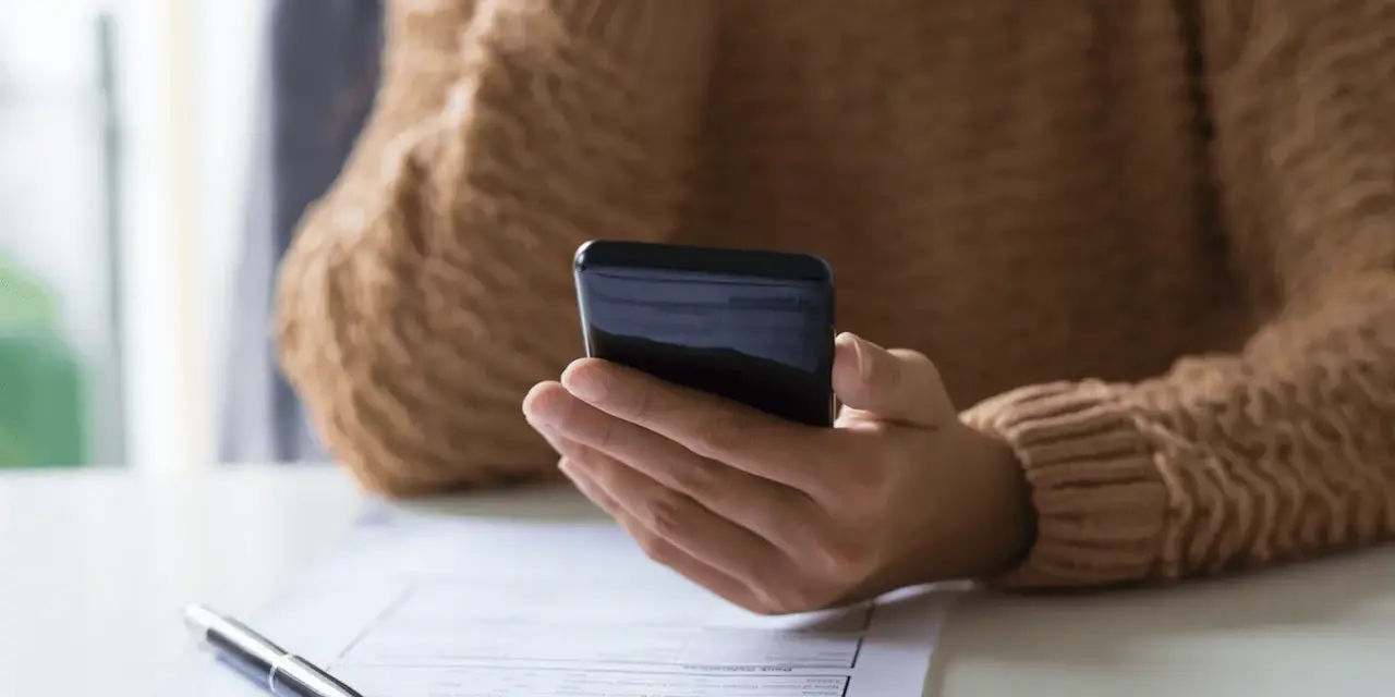 Persona revisa su teléfono móvil, modelo 035, mientras tiene un bolígrafo y un formulario de solicitud sobre la mesa.