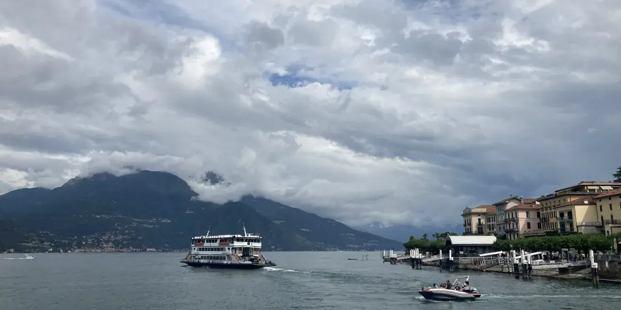 Promenada nad jeziorem Como, z górami w tle i statkiem wycieczkowym. W oddali widać urokliwe miasteczko.