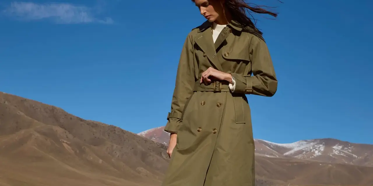 Mujer con un trench verde oliva, cinturón anudado, en un paisaje montañoso con cielo azul.