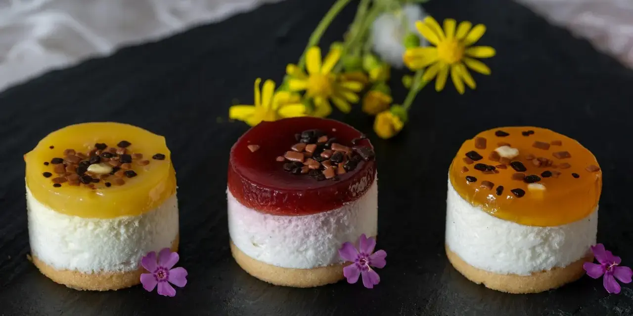 Efektowne ciasto bez pieczenia: trzy mini deserki z galaretką, kremem i owocami, ozdobione kwiatami i czekoladą.