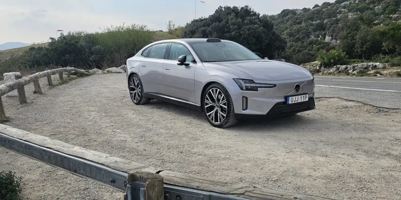 Srebrny samochód elektryczny zaparkowany na żwirowej drodze, otoczony bujną zieloną strefą i skalistymi wzgórzami.