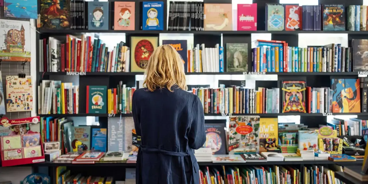Donna bionda in libreria, circondata da scaffali pieni di libri colorati. Un'atmosfera di lavoro e scoperta tra le pagine.