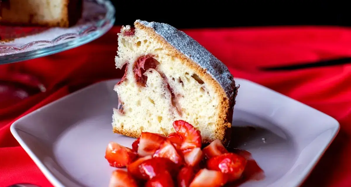 Un trozo de bizcocho de fresa esponjoso, espolvoreado con azúcar glas, servido con fresas frescas cortadas en un plato rosa.