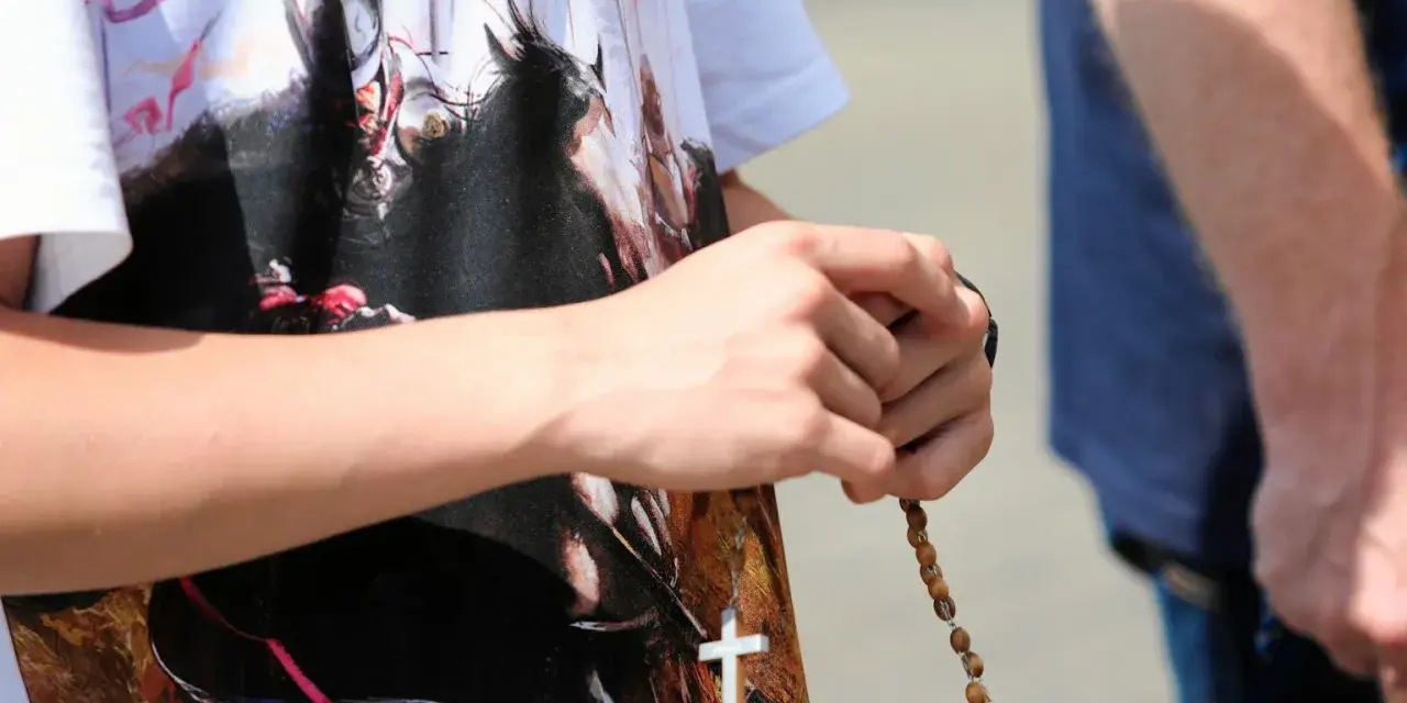 Dłoń trzyma paciorki różańca, a na koszulce widnieje biały krzyż. To znak modlitwy, żeby się wszystko ułożyło.