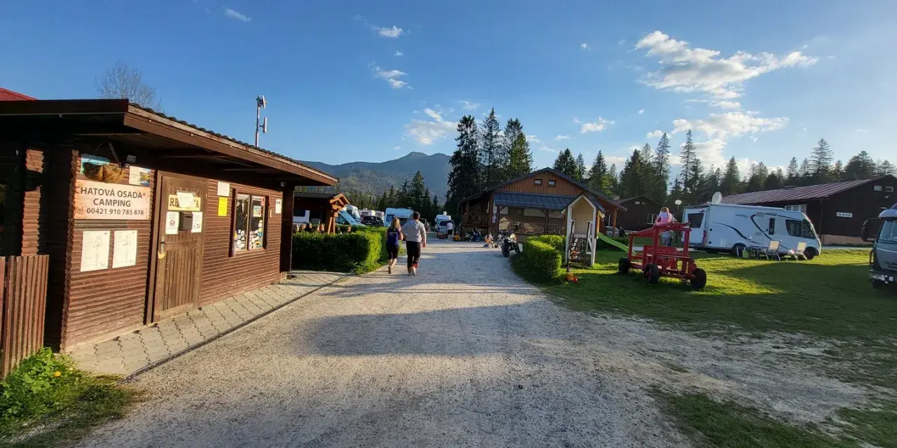 Słoneczny dzień na **camping Oravice**. Drewniany budynek recepcji, przyczepy kempingowe i zielone wzgórza w tle.