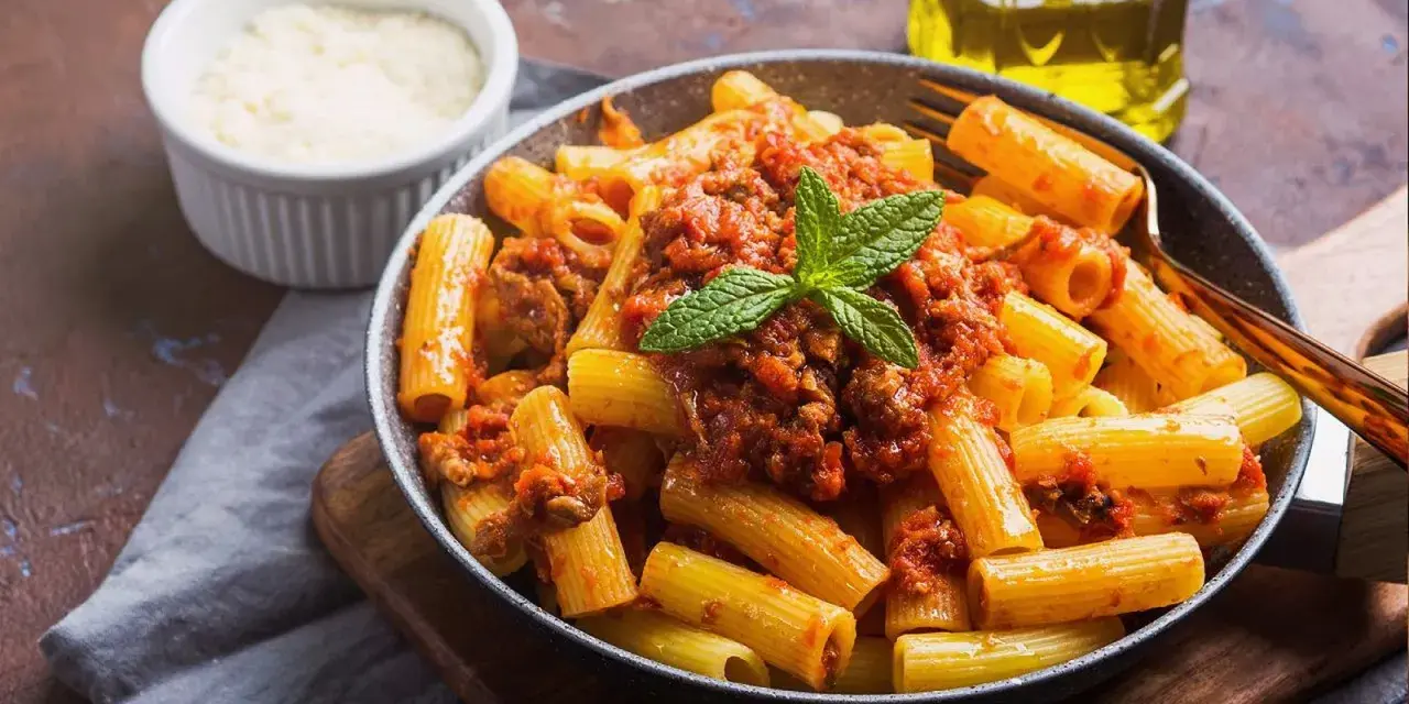 Un plato de macarrones con carne picada, cubiertos con salsa de tomate y decorados con menta. Al lado, un cuenco de queso rallado y una botella de aceite de oliva.