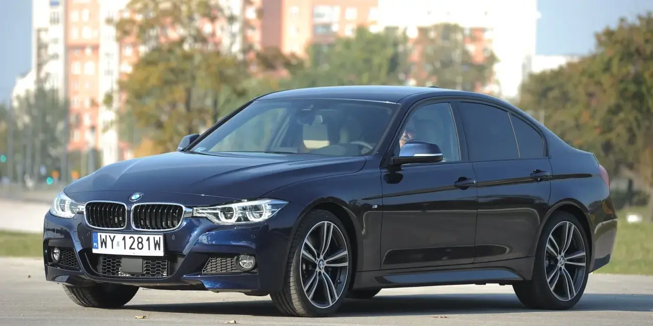 Ciemnoniebieskie BMW 318d z polskimi tablicami rejestracyjnymi stoi na tle budynków i drzew.