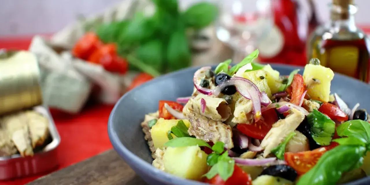 Un'insalata pantesca colorata con patate, pomodorini, olive, cipolla rossa e pesce, guarnita con basilico fresco.