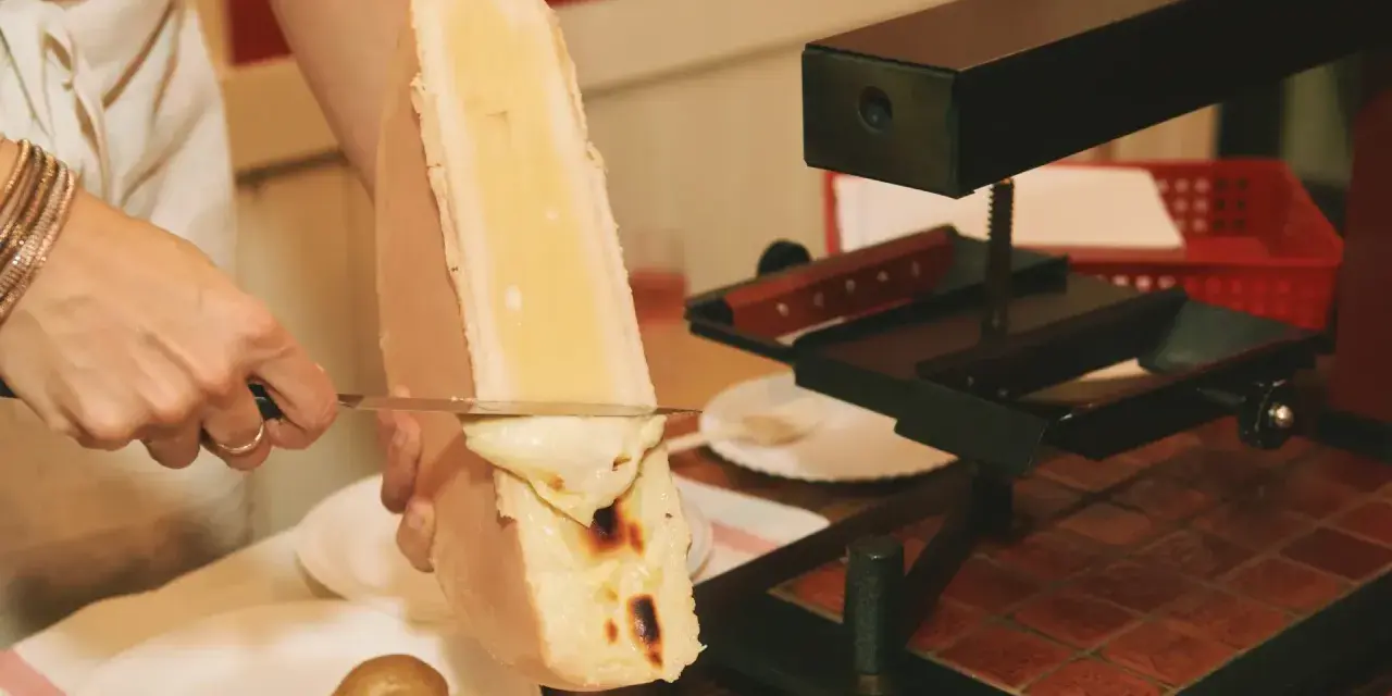 En person skrapar smält ost från en stor ostbit med en kniv. Perfekt för en mysig kväll med vin till raclette.