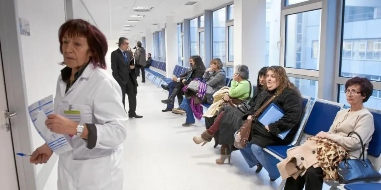 Doctora con bata blanca revisa papeles, mientras pacientes esperan en pasillo. Quizás pensando en cómo pedir excedencia estando de baja.
