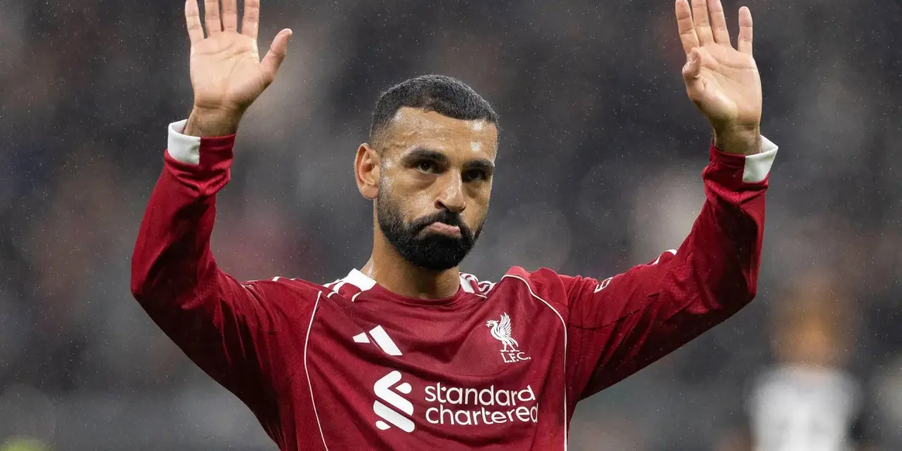 Mohamed Salah w czerwonej koszulce Liverpoolu, z uniesionymi dłońmi, pozdrawia kibiców. Widać logo klubu i sponsora.