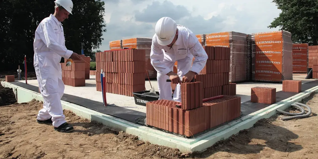 Dwóch budowlańców w białych kombinezonach układa cegły na fundamencie.