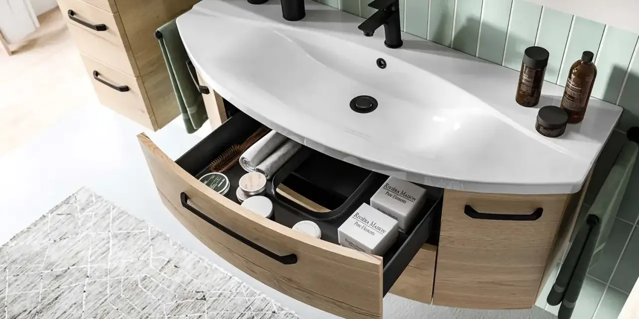 Aménagement salle de bain moderne avec lavabo blanc, robinetterie noire et tiroirs en bois clair organisés.