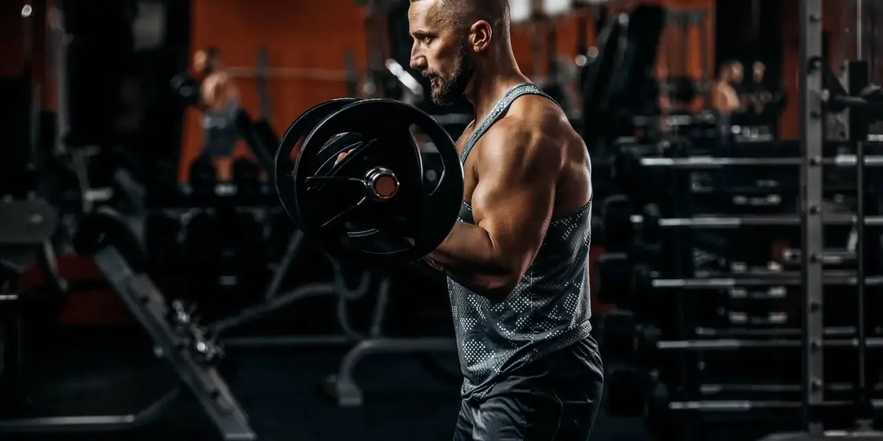 Mężczyzna ćwiczy biceps z ciężarem. W tle siłownia, sprzęt i inny ćwiczący.