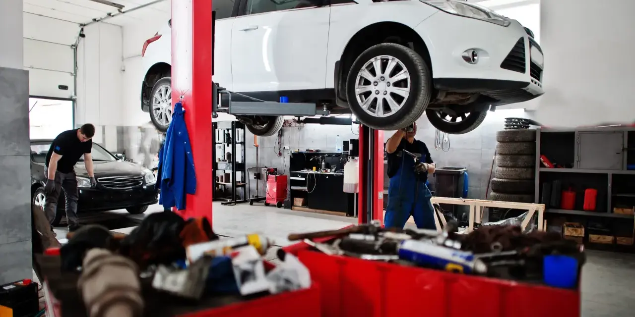 Warsztat samochodowy: biały samochód na podnośniku, mechanik w niebieskim kombinezonie.