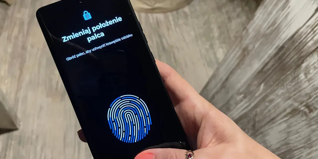Dłoń z telefonem, na ekranie skaner linii papilarnych i komunikat "Zmieniaj położenie palca". To jak próba ucieczki z cyfrowego więzienia, niczym **jailbreak**.