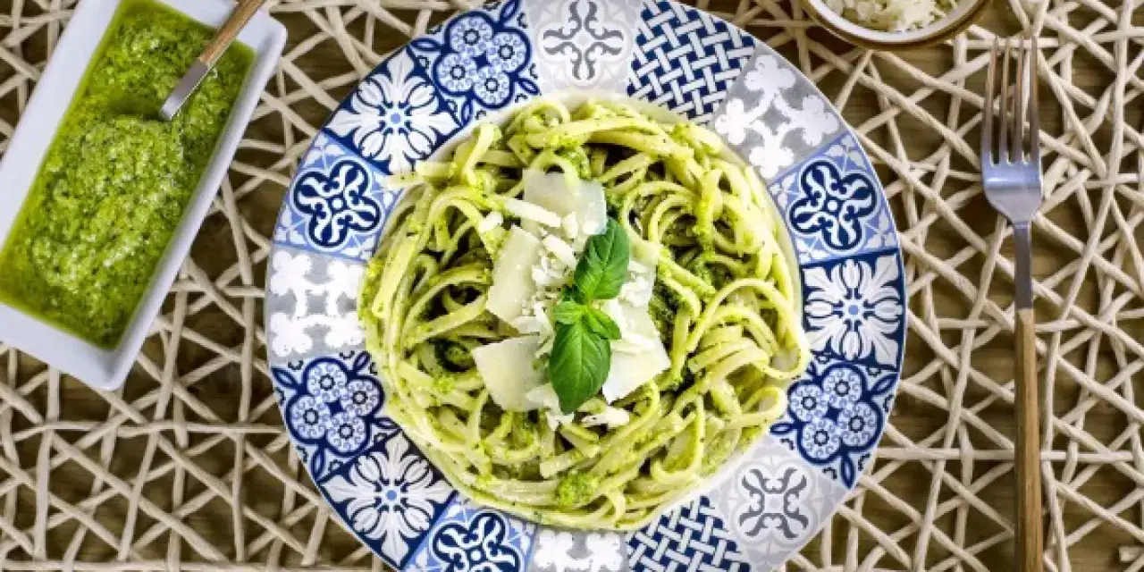 Pasta al pesto servida en plato decorado, con salsa de pesto y queso rallado al lado.