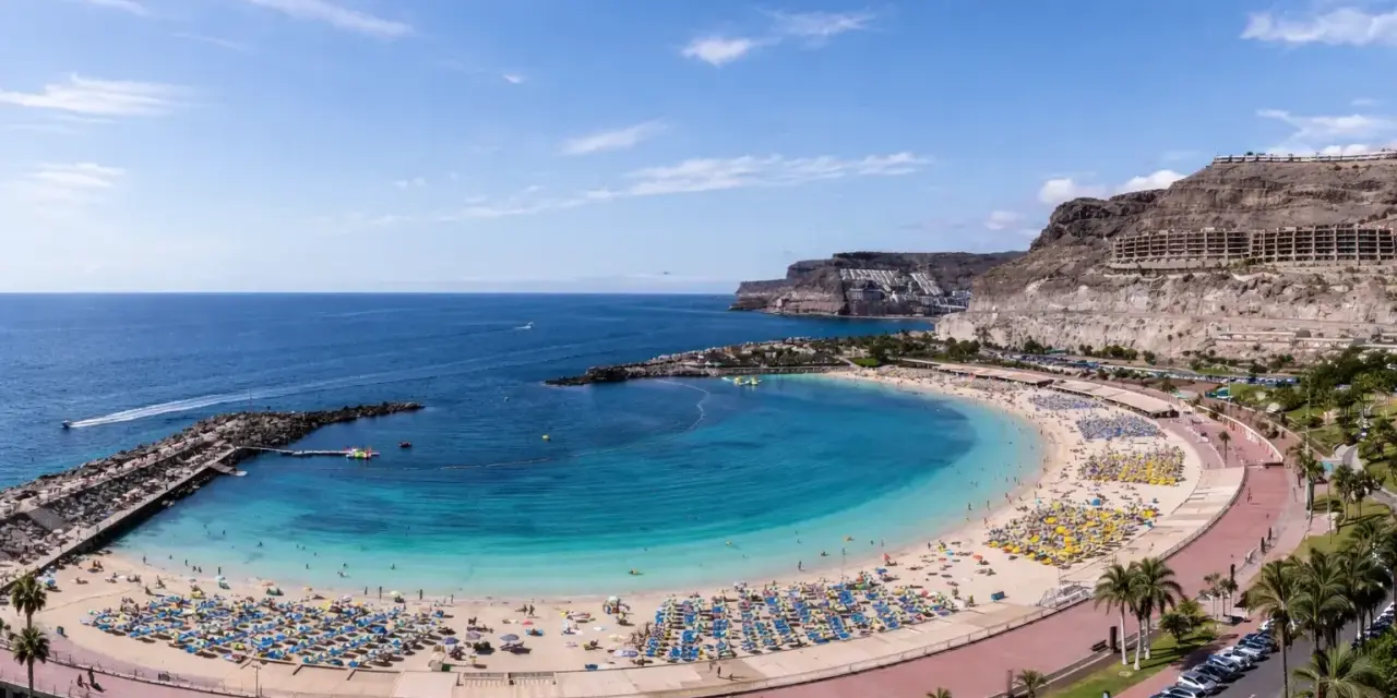 Słoneczna plaża z turkusową wodą i mnóstwem leżaków. W oddali klify i hotele, idealne gran canaria atrakcje dla każdego.