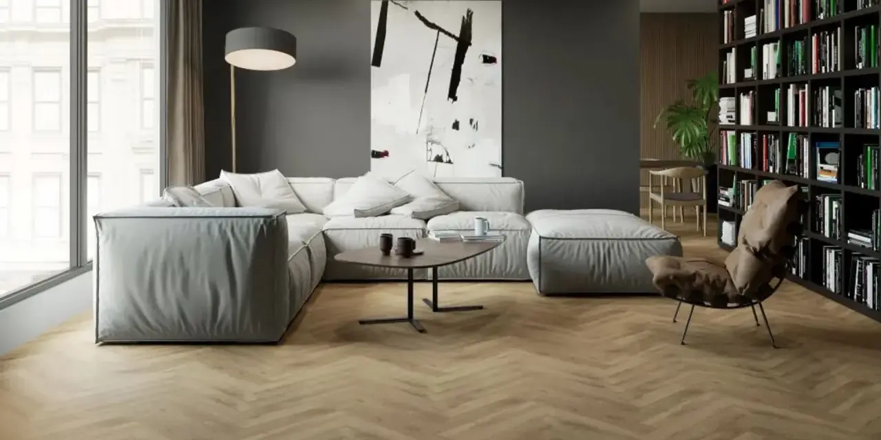 Nowoczesny salon z szarą kanapą i regałem pełnym książek. Podłoga w jodełkę imituje drewno, pokazując, jak dobrać płytki drewnopodobne do paneli.