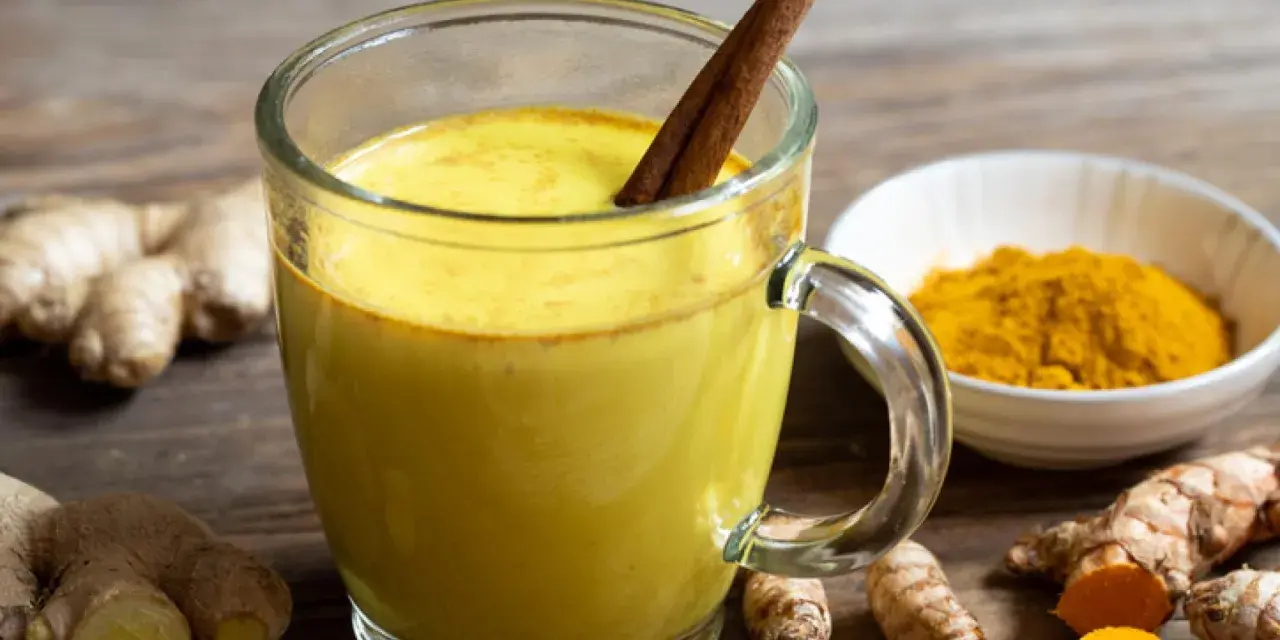 Taza de leche dorada con canela, rodeada de raíz de cúrcuma fresca y en polvo.