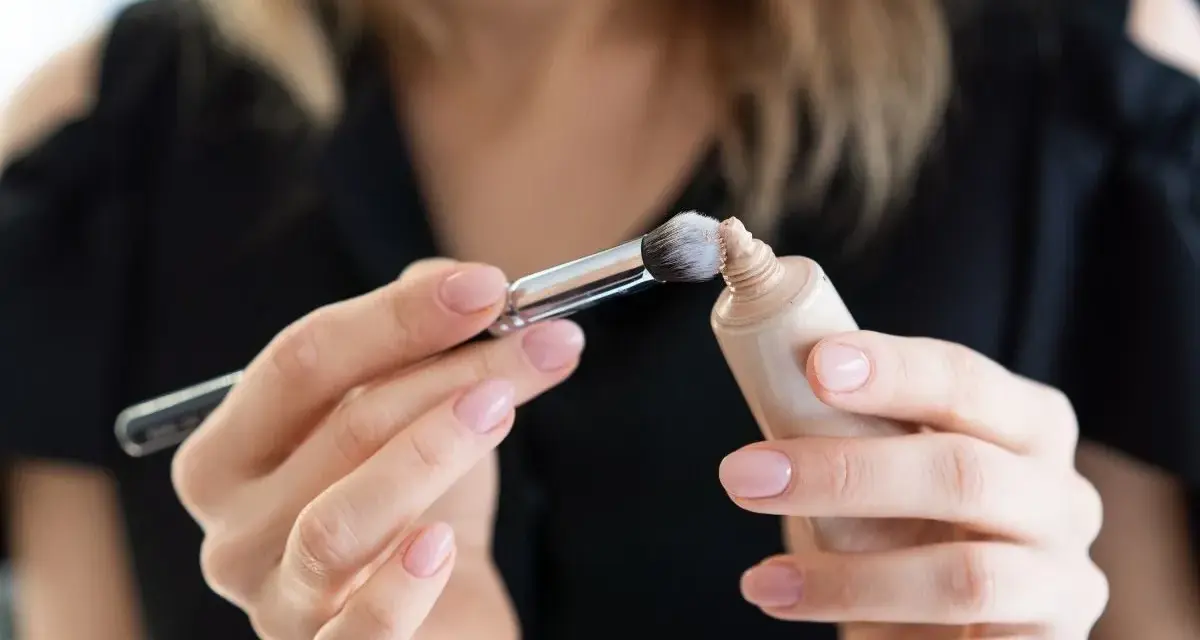 Dłonie z perfekcyjnym manicure nakładają pędzelkiem dr irena eris podkład rozświetlający na pociągłą tubkę.