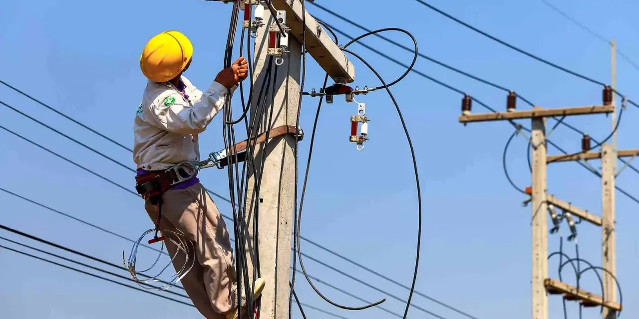 Elektryk w żółtym kasku pracuje na słupie energetycznym, podłączając przewody. Zastanawiasz się, ile kosztuje przyłącze elektryczne?