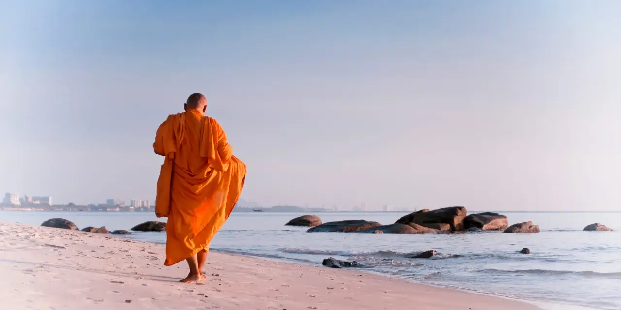 A modern day monk in an orange robe walks along a serene beach at sunrise, with calm ocean waves and distant city skyline.