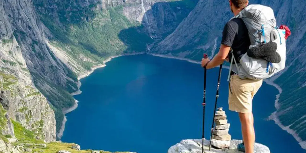 Un escursionista con zaino e bastoncini ammira un lago blu intenso circondato da montagne imponenti. Un'avventura di trekking montagna indimenticabile.