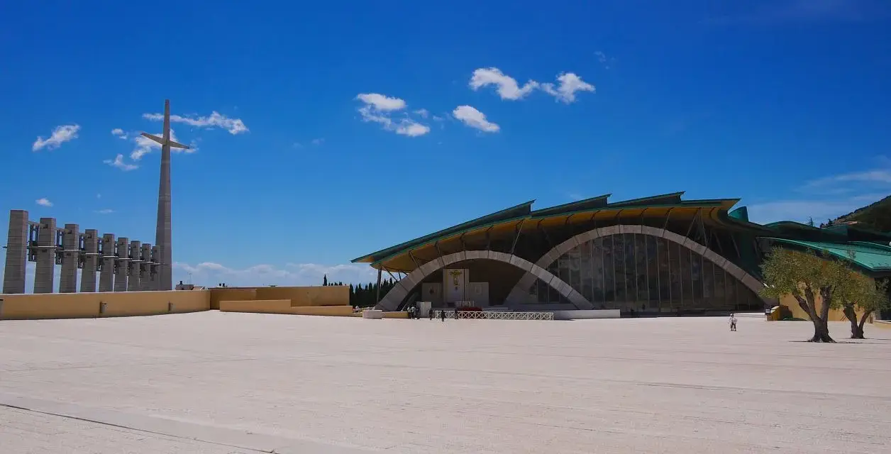 The modern church of San Giovanni Rotondo Padre Pio stands under a bright blue sky, with a tall cross and bell tower in the distance.