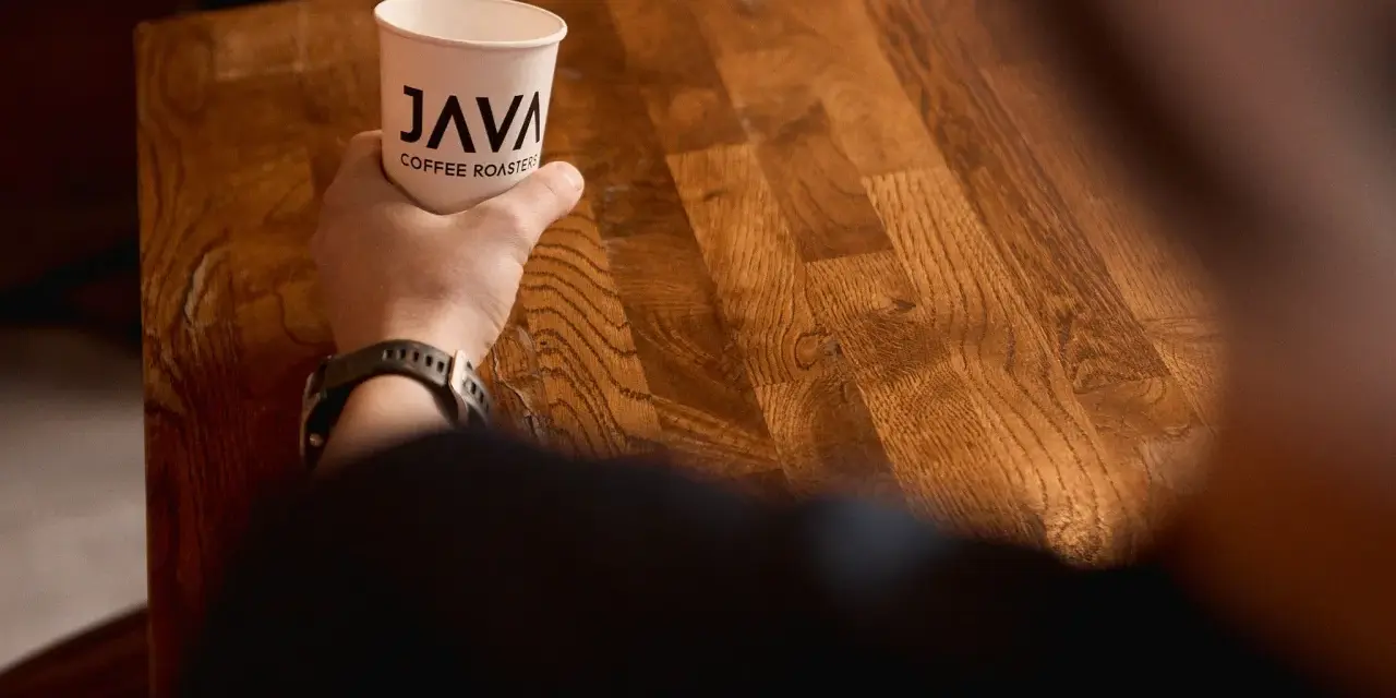 Dłoń trzyma kubek z logo "JAVA COFFEE ROASTERS", gotowa do degustacji świeżego batch brew na drewnianym stole.