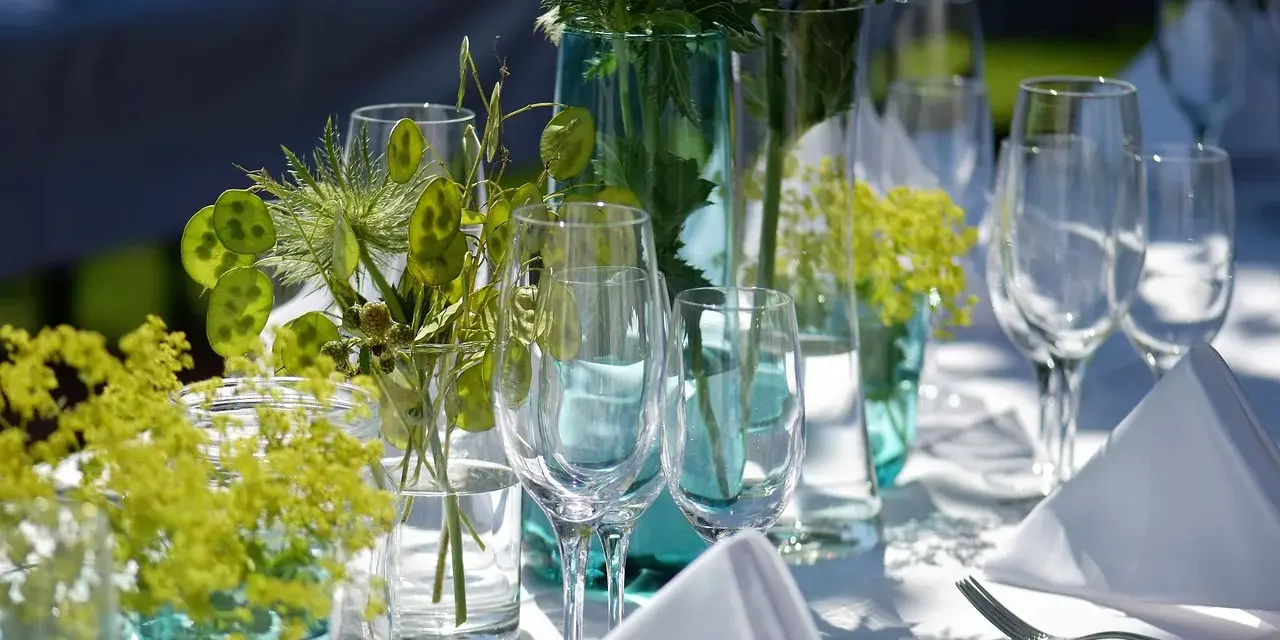 Eleganckie wesele w ogrodzie. Stół nakryty białym obrusem, ozdobiony zielonymi roślinami i kieliszkami.