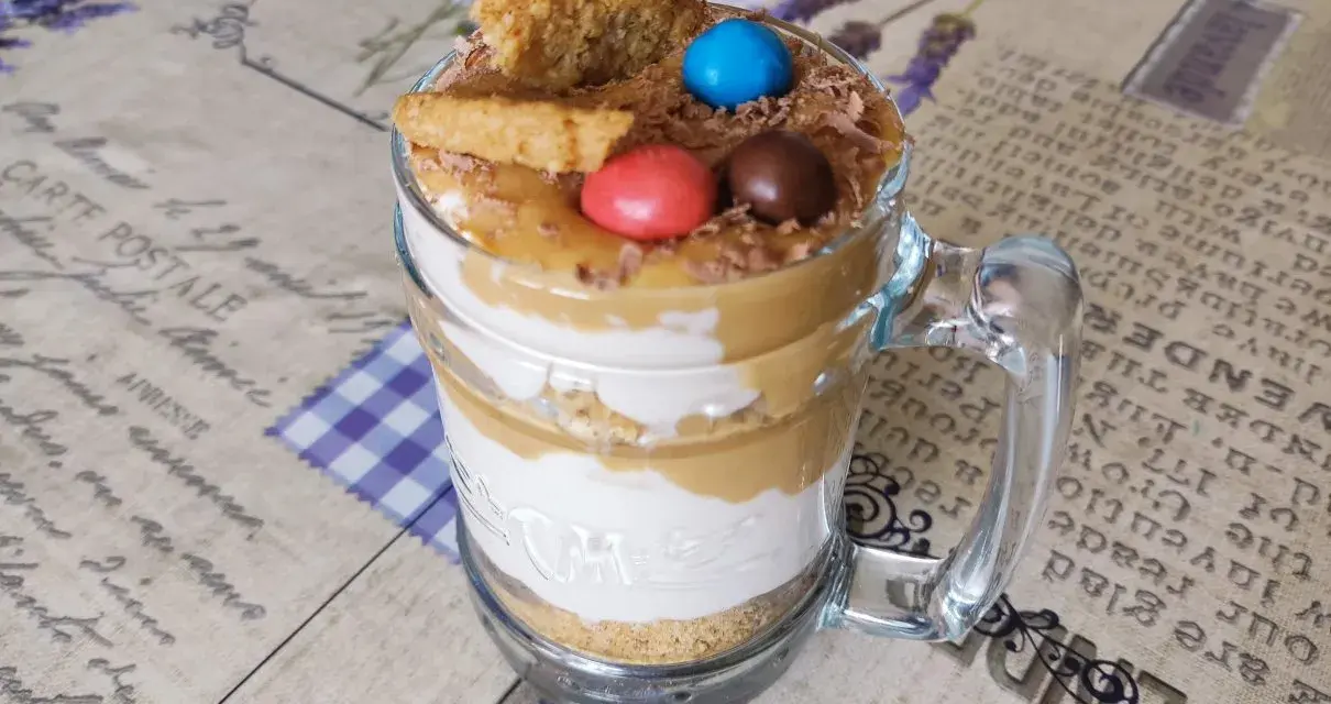 Warstwowy deser banoffee w szklance, ozdobiony ciasteczkiem, cukierkami i wiórkami czekolady.