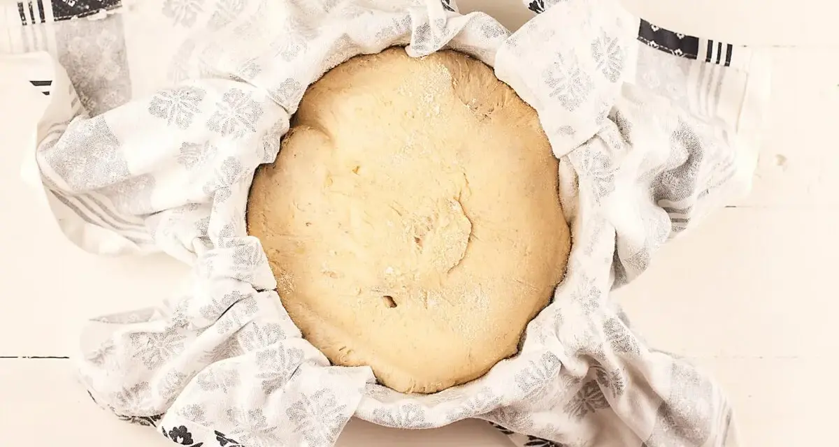 Masa de pan de patata cruda, cubierta con un paño blanco con estampado floral gris, lista para levar.