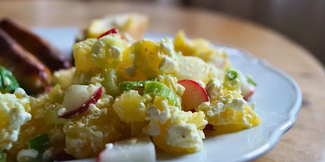 Kartoffelsalat mit Radieschen, Sellerie und Frischkäse auf einem Teller. Daneben liegen Würstchen.