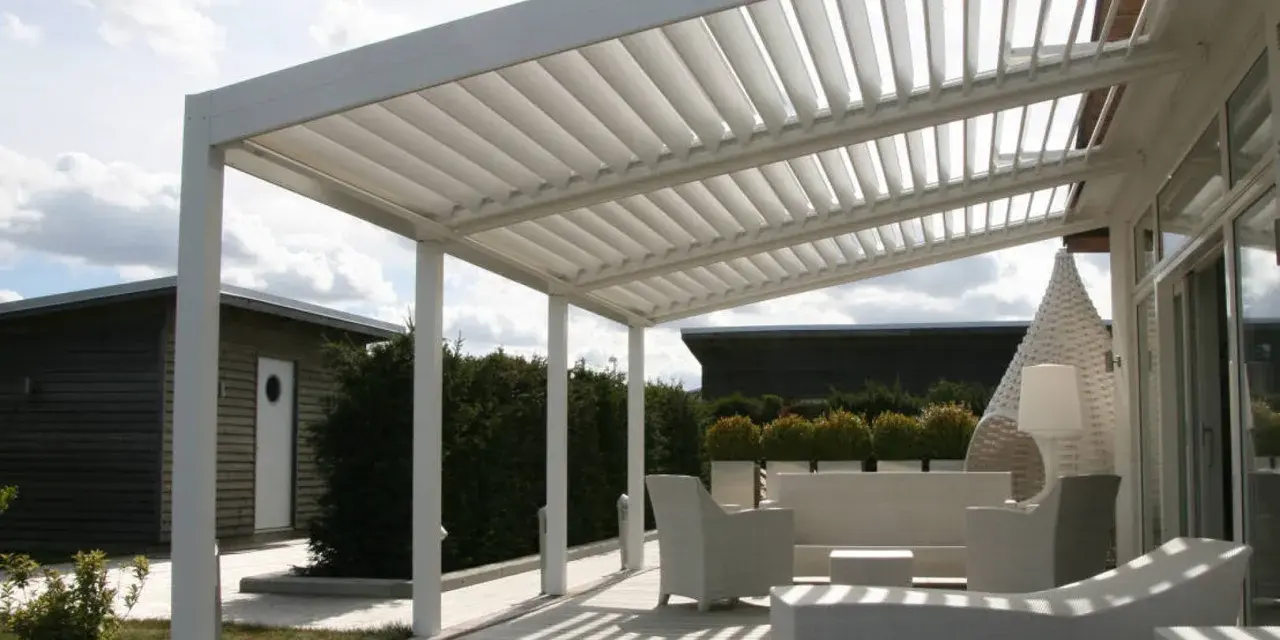 Un moderno porche de obra con techo de lamas ajustables, mobiliario de exterior blanco y un camino de madera que conduce a una cabaña.