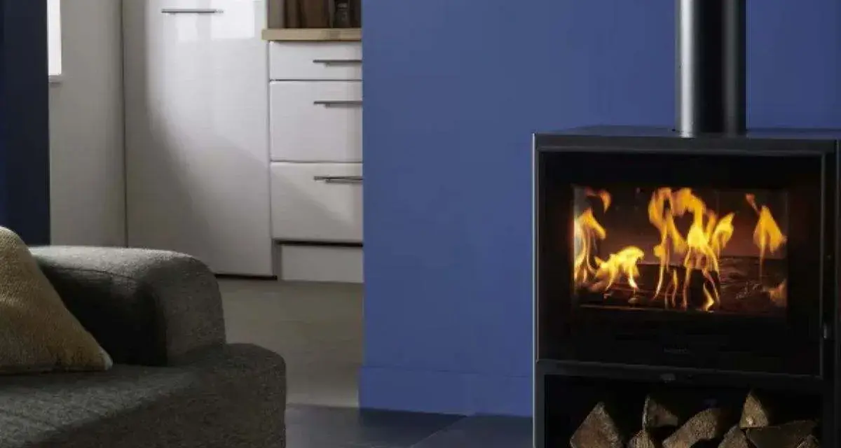 Estufa de leña moderna con fuego encendido y leña apilada, junto a un sofá y muebles de cocina.
