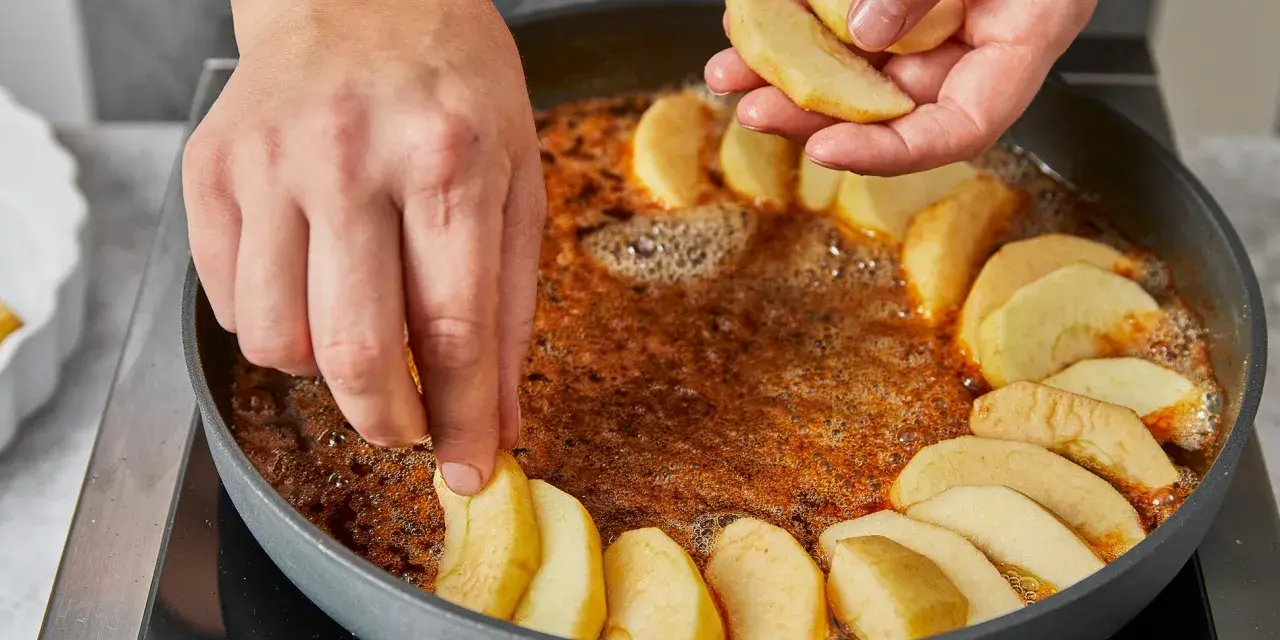 Hände legen Apfelschnitze in eine Pfanne mit karamellisierter Butter. Die Zubereitung der tarte tatin original beginnt.