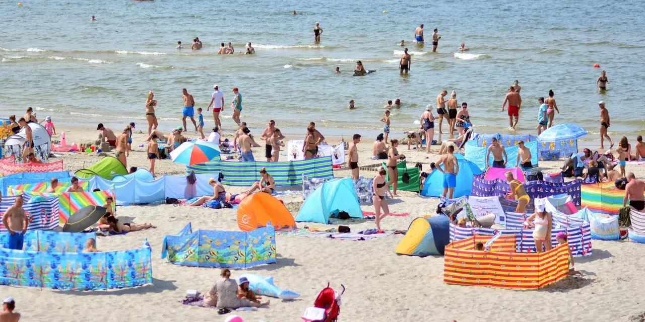 Wczasy pod gruszą na zatłoczonej plaży. Ludzie kąpią się w morzu, opalają na piasku i odpoczywają pod kolorowymi parawanami.