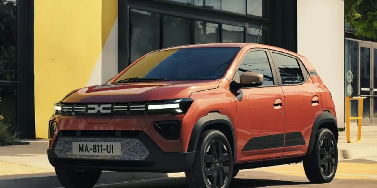 Pomarańczowy Dacia Spring stoi przed budynkiem z żółtą ścianą.