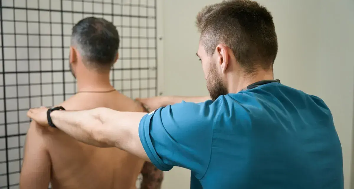 Therapeut führt krankengymnastik schulter Übungen durch. Ein Mann steht vor einem Gitter, während ein Therapeut seine Schultern berührt.