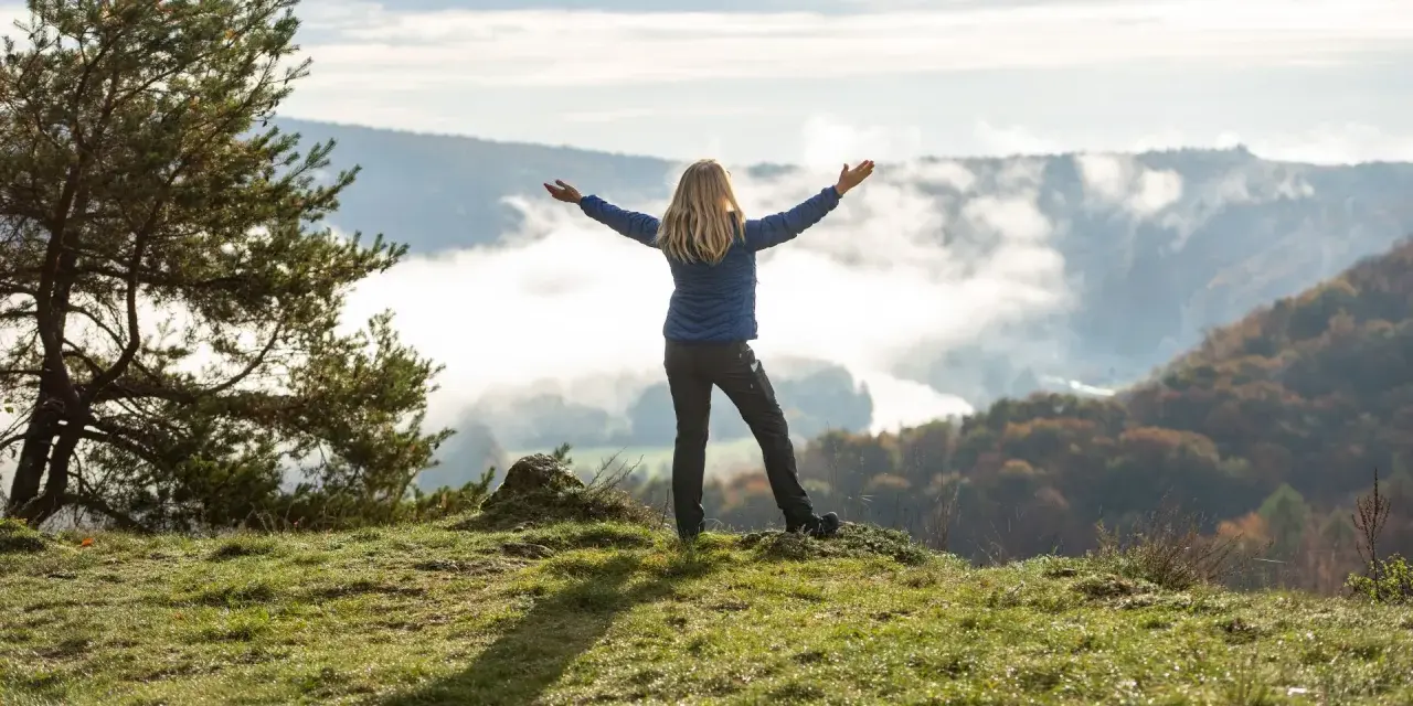 Frau mit ausgebreiteten Armen auf einem Hügel, umgeben von Nebel und Herbstfarben. Das Gefühl von Freiheit und die Bedeutung des Pilgerns werden hier spürbar.