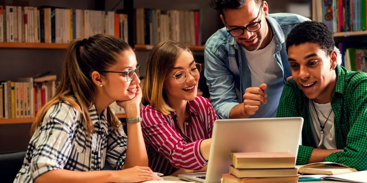 Vier Studierende lernen in der Bibliothek. Ein Mann, der wie ein seelsorger quereinsteiger wirkt, erklärt etwas am Laptop.