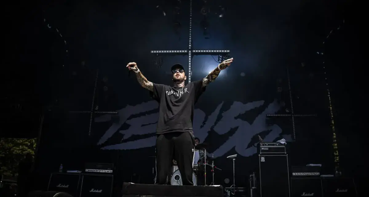 Un rappeur tatoué, casquette et lunettes de soleil, bras levés sur scène, devant un fond noir avec le logo "TEN 56".