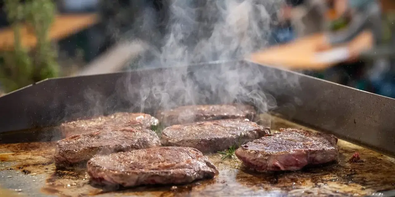 Mehrere saftige Rumpsteaks braten auf einer heißen Platte, Dampf steigt auf.