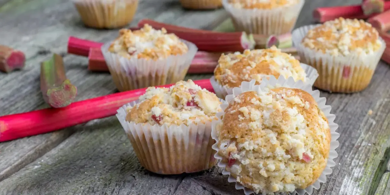 Pyszne muffinki z rabarbarem, posypane kruszonką, leżą na drewnianym blacie obok świeżych łodyg rabarbaru.