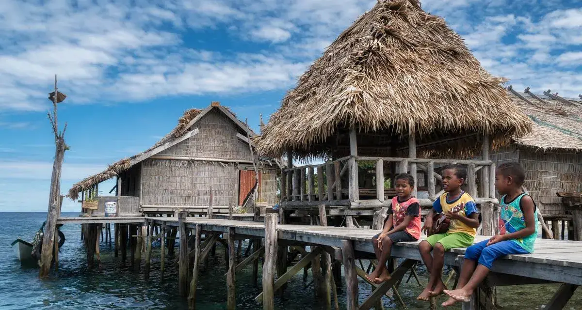 Trzech chłopców siedzi na drewnianym pomoście przy tradycyjnych chatach na wodzie w Raja Ampat.